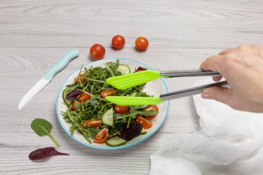 Taze domatesli salata, salatalık, roka, fesleğen, tabakta ıspanak. Paslanmaz çelik salata maşasıyla kadın eli. Sağlıklı sebze diyeti menüsü. Üst görünüm.