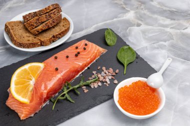 Çiğ somon balığı filetosu roka, fesleğen yaprağı, baharat ve limon dilimleri. Kırmızı havyarlı bir kase ve mutfak masasında çavdar ekmeği olan bir tabak. Üst görünüm.