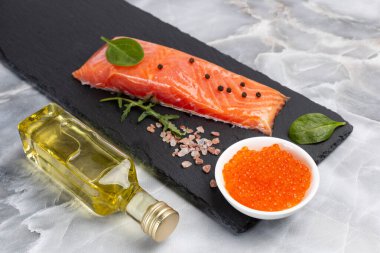 Çiğ somon balığı filetosu roka, fesleğen yaprağı, baharat ve dilimlenmiş limon. Kırmızı havyarlı bir kase ve mutfak masasında bir şişe yağ. Üst görünüm.