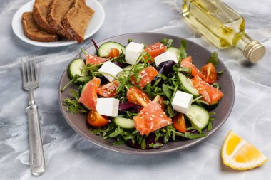 Dilimlenmiş somon balıklı salata, beyaz peynirli feta, taze domates, salatalık, roka, fesleğen, çatallı ıspanak. Bir dilim limon, bir tabak çavdar ekmeği ve bir şişe zeytinyağı. Üst görünüm.