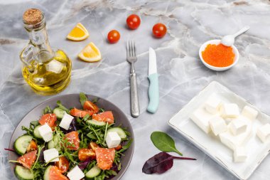 Dilimlenmiş somon balıklı salata, beyaz peynirli feta ve tabakta taze sebze. Kırmızı havyarlı kase, dilimlenmiş limon, bir şişe yağ ve mutfak masasında peynirli bir tabak. Üst görünüm.