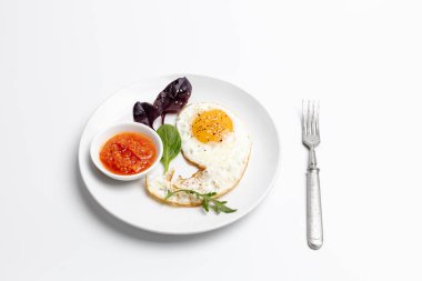 Beyaz arka planda kızarmış yumurta, kırmızı sos ve fesleğen yapraklı beyaz porselen tabak. Üst görünüm.