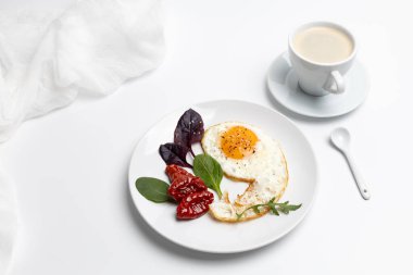 Karabiberli sahanda yumurta, susam, kurutulmuş domates, roka ve beyaz tabakta fesleğen. Beyaz arka planda bir fincan sade kahve. Üst görünüm.