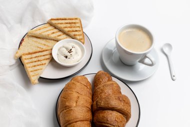 Kahvaltıda peynirli tost, kruvasan, beyaz porselen bir fincan sade kahve ve beyaz arka planda bir kaşık. Üst görünüm.