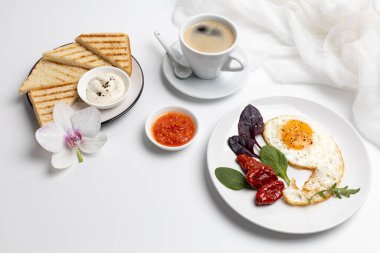 Tavada yumurta, kurutulmuş domates ve fesleğen yaprağı. Tost tabağı ve bir orkide çiçeği. Kırmızı domates sosu ve peynir soslu kaseler. Beyaz arka planda bir fincan sade kahve..