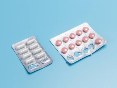 Beyaz kapsüllü ilaç paketleri ve mavi arka planda pembe haplar. Üst görünüm.