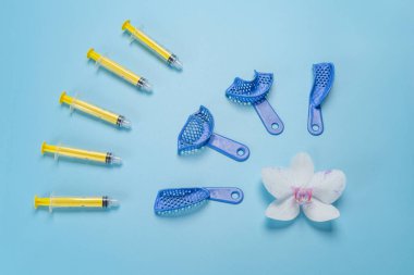 Set of dental flexible impression trays with plastic syringes and an orchid flower on the blue background. Top view. clipart