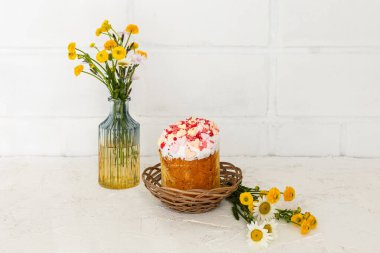 Hasır sepetlerde sarı Ranunculus asiaticus ve cam vazoda Paskalya pastası. Arka planda beyaz duvar.