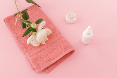 Yüz ve vücut bakımı ürünleri. Şakayık çiçekli yumuşak havlu, yüz serumu ve pembe arka planda yüz kremi. Spa ve vücut bakımı seti. Üst görünüm.