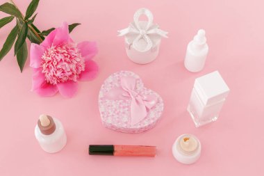 Hediye kutuları, yüz serumu, ruj, yüz kremi, bir şişe parfüm ve pembe arka planda şakayık çiçeği. Kadın kozmetik ve aksesuarları. Üst görünüm.