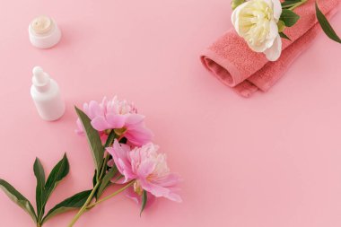 Yüz ve vücut bakımı ürünleri. Şakayık çiçekli yumuşak havlu, yüz serumu ve pembe arka planda yüz kremi. Spa ve vücut bakımı seti. Üst görünüm.