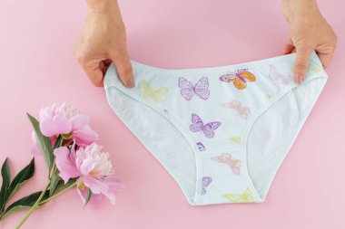 Kadınların elleri pembe arka planda şakayık çiçekli bikinili pamuk külot tutuyor. En iyi günlük iç çamaşırları. Üst görünüm.