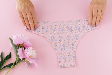 Kadınların elleri, pembe arka planda, üzerinde şakayık çiçekleri olan minimalist tanga tarzı pamuk külotları tutuyor. En iyi günlük iç çamaşırları. Üst görünüm.