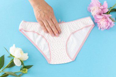 Mavi arka planda, üzerinde şakayık çiçekleri olan bikinili pamuk külot tutan bir kadın eli. En iyi günlük iç çamaşırları. Üst görünüm.