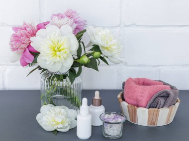 Yüz ve vücut bakımı ürünleri. Yüz serumu şişeleri, deniz tuzu ve yumuşak havlularla dolu bir kase ahşap sepette şakayık çiçekleri. Spa ve vücut bakımı konsepti. Üst görünüm.