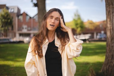 Parktaki güçlü baş ağrısı olan çekici kadın. Güzel bir esmerin portresi şakaklarına dokunuyor. Stres ya da acı hissediyor. Stres veya baş ağrısı konsepti. Stres yüzünden. Negatif insani duygu.