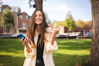 Portre fotoğrafı gülümseyen genç kız cep telefonuyla internette geziniyor. Günlük kıyafet tişörtü giyiyor ve kazanan gibi hareket ediyor. Esmer kadın mutlu görünüyor, zaferi, zaferi, başarıyı kutluyor..