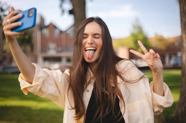 Parkta yürürken cep telefonuyla selfie çeken gülümseyen esmer bir kadın resmi. Barış jesti yap, iki parmağını göster, mutlu görün, gülümse, dilini çıkar, gözlerini kapat..