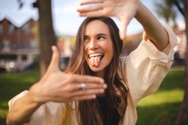 Çekici, neşeli esmer kız dışarıda güzel bir dinlenme tatili görüntüsü gösterirken eğleniyor. Çerçeveyi arayan kız parmaklarıyla, diliyle bir kare yapıyor ve bakıyor..