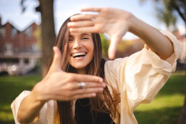 Çekici, neşeli esmer kız dışarıda güzel bir dinlenme tatili görüntüsü gösterirken eğleniyor. Kız çerçeveyi arıyor parmaklarıyla kare yapıyor ve bakıyor.