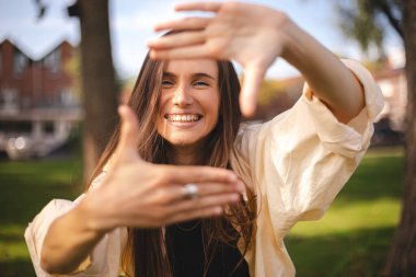 Çekici, neşeli esmer kız dışarıda güzel bir dinlenme tatili görüntüsü gösterirken eğleniyor. Kız çerçeveyi arıyor parmaklarıyla kare yapıyor ve bakıyor.