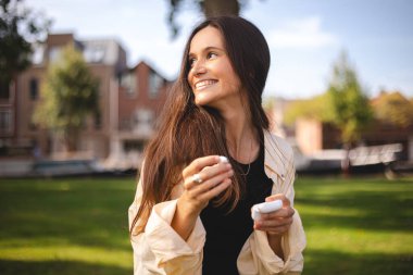 Esmer kadın kulaklıklarına kablosuz kulaklık takıyor. Kadın eli iki beyaz kablosuz bluetooth kulaklık tutuyor. Kız çantasını tut ve kulaklığını tak. Mutlu gülen kız müzik dinle, sesli kitap oku