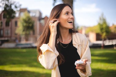 Esmer kadın kulaklıklarına kablosuz kulaklık takıyor. Kadın eli iki beyaz kablosuz bluetooth kulaklık tutuyor. Kız çantasını tut ve kulaklığını tak. Mutlu gülen kız müzik dinle, sesli kitap oku