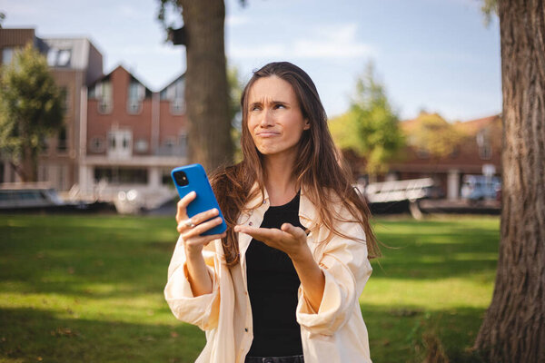 Beautiful upset woman walking outdoor in park. Sad angry female having talking conversation with mobile phone, yelling. Screaming woman looking phone feels shock upset worries bad news problems.