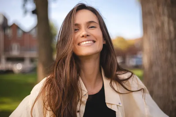 Gömlekli güzel bir kadın, tebessüm ediyor ve kırsal kesimde kameraya bakıyor. Gülümseyen esmer bir kızın açık hava portresi. Mutlu neşeli kız yeşil şehre gülüyor, moda gibi görünüyor ve dişlek gülüşünü sergiliyor.