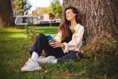 Parkta ağacın altında oturan bir kadın, gömlek, bluz, spor ayakkabı ve kot pantolon, cep telefonu ve kulaklık takıyor. Telefonunda müzik, podcast veya radyo açıyor ve kulaklıkla dinliyor..