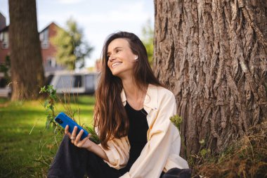 Kablosuz kulaklıklı ve cep telefonlu müzik, podcast ya da parktaki ağacın altında oturan sesli kitap dinleyen genç bir kadın. Düşünceli, gülen kadın rüyası. Mutlu duygular.