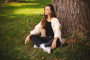 Esmer saçlı, müzik, mantra, ses kitabı ya da podcast dinleyen, gözleri ağacın altında olan genç, hoş görünümlü bir kadın. Kırsal alan. Kulaklıklı güzel kız meditasyon yapıyor..