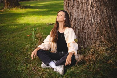 Esmer saçlı, müzik, mantra, ses kitabı ya da podcast dinleyen, gözleri ağacın altında olan genç, hoş görünümlü bir kadın. Kırsal alan. Kulaklıklı güzel kız meditasyon yapıyor..