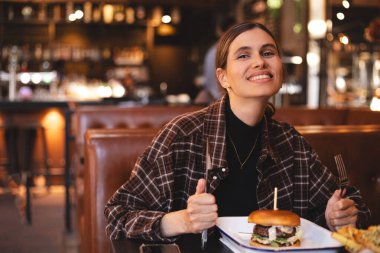 Restoranda hamburger yiyen bir kadın, çatal ve bıçakla hazırlanmış. Hile yemeği, fast food. Fast food restoranındaki kafede oturan genç ve aç bir kadının portresi. Sağlıksız yemek..