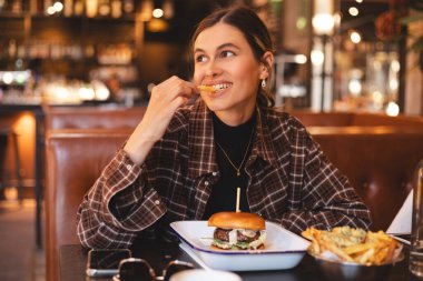 Fast food aşığı. Hamburger yiyen, patates yiyen, patates yiyen, gözlerini kapayan, restoranda oturan mutlu bir kadın. Elinde patates kızartması, tabakta burger ve lezzetli bir kadın. Hile yemekleri..