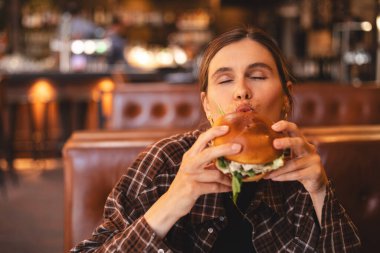 Close-up portrait of a woman in a fast food restaurant eating a hamburger. Hungry woman eating a burger sitting in cafe. Girl bite bbq burger, look joyful. Cheat meal day concept. clipart