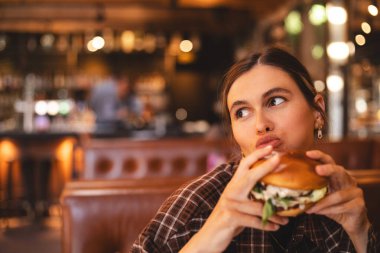 Fast food restoranında hamburger yiyen bir kadının yakın plan portresi. Kafede oturup hamburger yiyen aç bir kadın. Kız BBC hamburgeri yer, neşeli görün. Yemek günü konseptini aldatma.