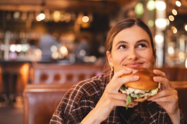 Fast food restoranında hamburger yiyen bir kadının yakın plan portresi. Kafede oturup hamburger yiyen aç bir kadın. Kız BBC hamburgeri yer, neşeli görün. Yemek günü konseptini aldatma.