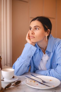 Young woman sitting at table after finishing a meal, wearing casual blue shirt, white top, look sad and disappeared. Table set with empty plates, cutlery, leftovers. Girl was not full. Hold her head. clipart