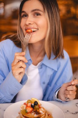 Çatalla çilek yiyen mutlu bir kadın, bir restoranın içinde meyveli krep ve akçaağaç şurubu yiyen ve yanına bakan bir kadın. Sarı saçlı mutlu kızı kapat ve kafede eğlen. Bıçak, çatal.