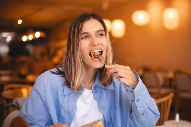 Kafede çatallı leziz krep yiyip onu yiyen mutlu bir kadın. Oburluk ve yeme bozukluğu kavramı. Kapalı tişört ve beyaz tişört giyen kız. Kapalı gözler.