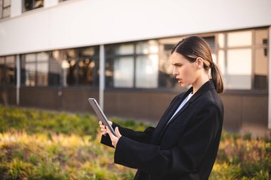 Şüpheli iş kadını elinde dijital tablet tutarken sinirli görünüyor, kafası karışık. At kuyruklu kadın, ofis binasının yanında duran spor ceket giyin. Tablete şaşırmış bak.