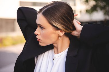 Sarışın bir iş kadınının yakın plan portresi. İş merkezi binasının yanında duran bir kadın. Tarz saç stili ve küpeleriyle yan tarafa bakıyor. Kız saç düzeltir, at kuyruğu yapar..