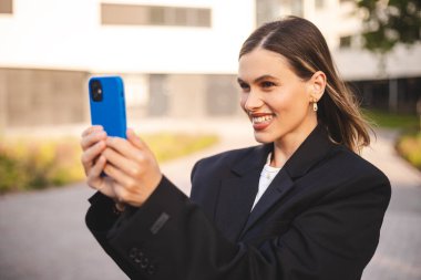 Video çağrısı, fotoğraf çek. Güzel bir iş kadını video görüşmesi yapıyor. Ofis binasının yanındaki caddede akıllı telefonuyla fotoğraf çekiyor. Mutlu iş kadını gülümsemesi tut cep telefonunu.