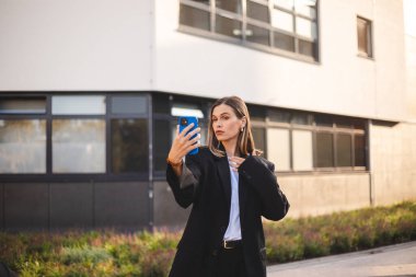 İş dünyasından ciddi bir kadın modern ofis binasına yaslanıyor. Ceket giymiş bir iş kadını telefonuyla fotoğraf çekiyor.. 