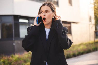 Akıllı telefondan konuşan iş kadını şok oldu. Kızgın genç bir kadın cep telefonuyla konuşuyor, kötü haberleri telefonla duyuyor, kızgın ve korkmuş görünüyor, ofisin yanında duran ağzı açık..