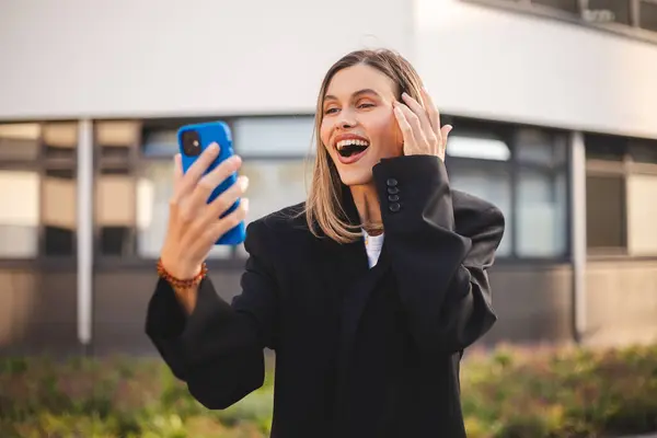 Dışarıyı aramak için cep telefonu kullanan güzel bir kadın. Ofisin yakınındaki bir caddede gülümseyen iş kızı akıllı telefon ekranı gibi görünüyor. Mutlu, gülümseyen bir kadın akıllı telefondan sohbet ediyor ceket giyiyor, konuşuyor..