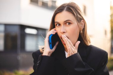 İş kadını ofisin yakınında telefonla konuşuyor, şok oldu. Sarışın kadın akıllı telefonunu kullanıyor. Şok edici bir haber. Ağzını elleriyle kapatmış. Dedikoducu kız. Kadınlar ilginç konuşmalar yapar..