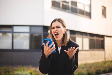 Siyah ceketli kızgın genç iş kadını ofisin dışında duruyor sesli mesajları kaydediyor ve agresif bir yüz ifadesiyle çığlık atıyor. Sinirli kız telefonla konuşuyor, yemin ediyor. Hayal kırıklığına uğramış kısa saçlı kadın..