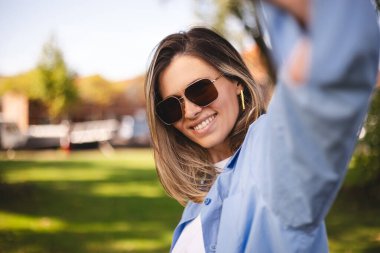 Gülümseyen genç sarışın kadın parkta yürürken elinde telefonuyla selfie çekiyor. Kız telefonuyla görüntülü arama yapıyor, mutlu görünüyor ve gülümsüyor. Güneş gözlüğü ve gömlek takan kadınlar selfie çeker..